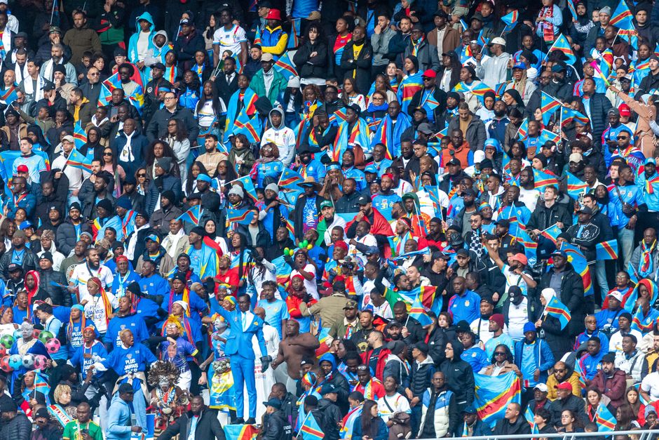 Packed stadium with diverse crowd waving Congolese flags, celebrating unity.