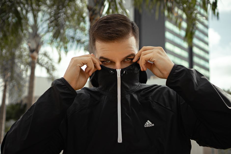 A man in a black jacket covers his face outdoors with modern architecture and palm trees.