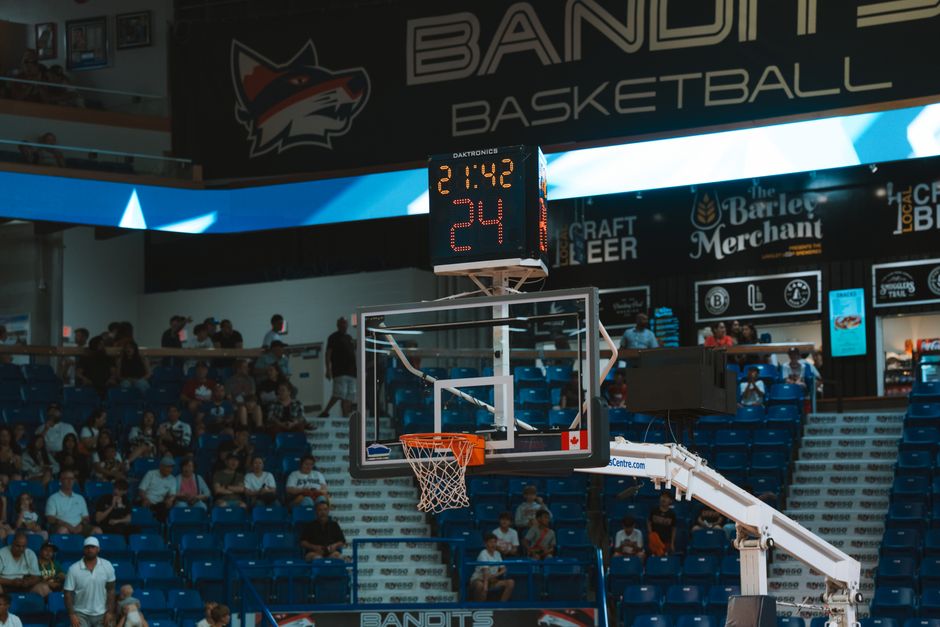 Close-up of a basketball hoop inside a stadium with a scoreboard and audience in the background.