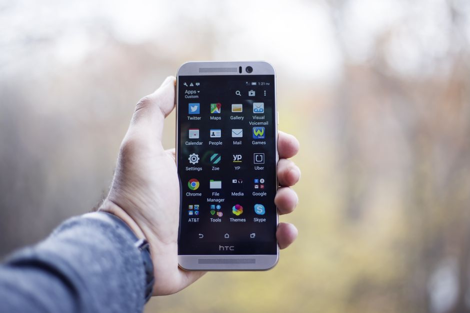 A close-up of a hand holding an HTC smartphone with visible apps on the screen, in an outdoor setting.