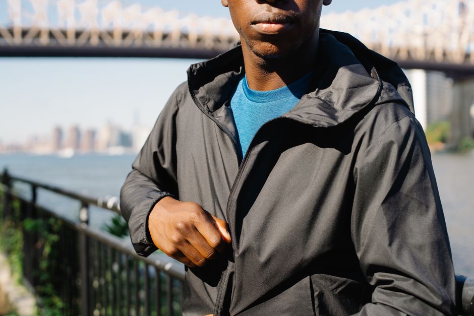 Stylish black man zips up windbreaker jacket near an iconic bridge on a sunny day.